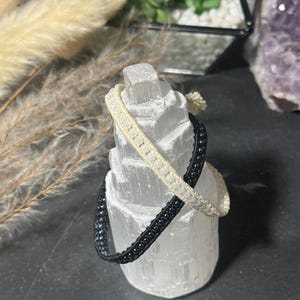 May include: Two macrame bracelets, one black and one cream-colored, are displayed on a white selenite crystal. The bracelets are intricately woven with small beads. The background includes dried plants and a small succulent.