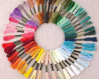 Puede incluir: Una disposición circular de hilos de bordar en una variedad de colores. Los hilos muestran una amplia gama de tonos, desde rojos y naranjas vibrantes hasta azules y verdes fríos, con tonos neutros intercalados. Cada madeja está etiquetada.