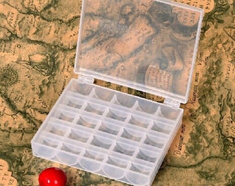 Op de afbeelding: Een doorzichtige plastic organizer met een scharnierend deksel, verdeeld in talrijke kleine compartimenten. Het deksel heeft een vintage kaartontwerp. Een klein, rood, rond object ligt naast de organizer. De organizer staat op een achtergrond met kaartthema.