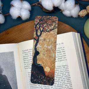 Puede incluir: Un marcapáginas con una ilustración de acuarela de una mujer sentada bajo un árbol leyendo un libro. El fondo es una puesta de sol con un cielo dorado y un sol rojo. El marcapáginas tiene el texto "Jotun Keatt" escrito en él.