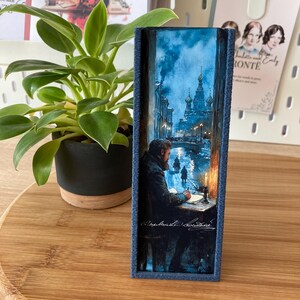 May include: A rectangular, blue-framed art piece depicting a man writing at a desk in a snowy, nighttime cityscape. The artwork features a church with onion domes and figures walking on a bridge. The frame has a textured, stitched appearance.