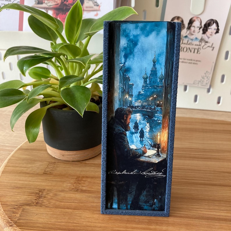 May include: A rectangular, blue-framed art piece depicting a man writing at a desk in a snowy, nighttime cityscape. The artwork features a church with onion domes and figures walking on a bridge. The frame has a textured, stitched appearance.