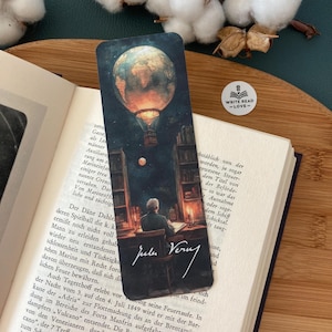 Könnte beinhalten: Ein Lesezeichen mit einer Vintage-Illustration eines Heißluftballons mit Globusdesign, einem Mann, der an einem Schreibtisch schreibt, und der Unterschrift "Jules Verne". Das Lesezeichen befindet sich in einem offenen Buch.