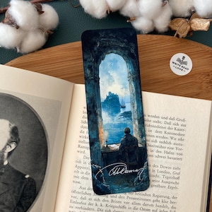 Könnte beinhalten: Lesezeichen mit einem Gemälde einer Person an einem Schreibtisch mit Blick auf eine Seelandschaft mit einer Burg. Das Lesezeichen hat hauptsächlich dunkelblaue und graue Farben, mit dem Text "WRITE READ -LOVE-" auf einem Aufkleber.