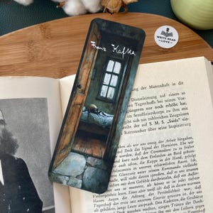 Könnte beinhalten: Ein Lesezeichen mit einer dunklen, stimmungsvollen Illustration eines Raumes mit einer angelehnten Tür, einem Bett und einer Spinne. Das Lesezeichen ist mit dem Text "Franz Kafka" in weißer Schrift bedruckt.
