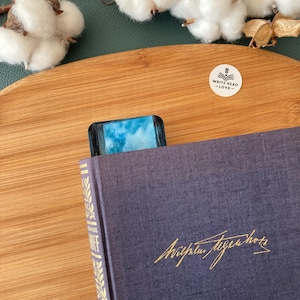May include: A blue and white art print bookmark is inserted into a dark blue book. The bookmark has a sky-like design. The book has gold lettering and a gold spine. A sticker with the words "WRITE READ -LOVE-" is on a wooden surface.