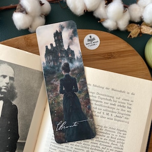 Puede incluir: Un marcapáginas con una ilustración de acuarela de una mujer con un vestido negro caminando hacia una gran casa de estilo gótico oscuro con humo saliendo de la chimenea. El texto "Bronte" está escrito en cursiva blanca en la parte inferior del marcapáginas.