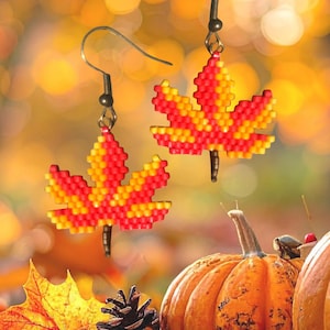 May include: A pair of dangle earrings featuring a beaded maple leaf design in vibrant shades of red and yellow. Each leaf is constructed from small, square-shaped beads and hangs from a dark metal hook, creating a distinct autumn-inspired accessory.