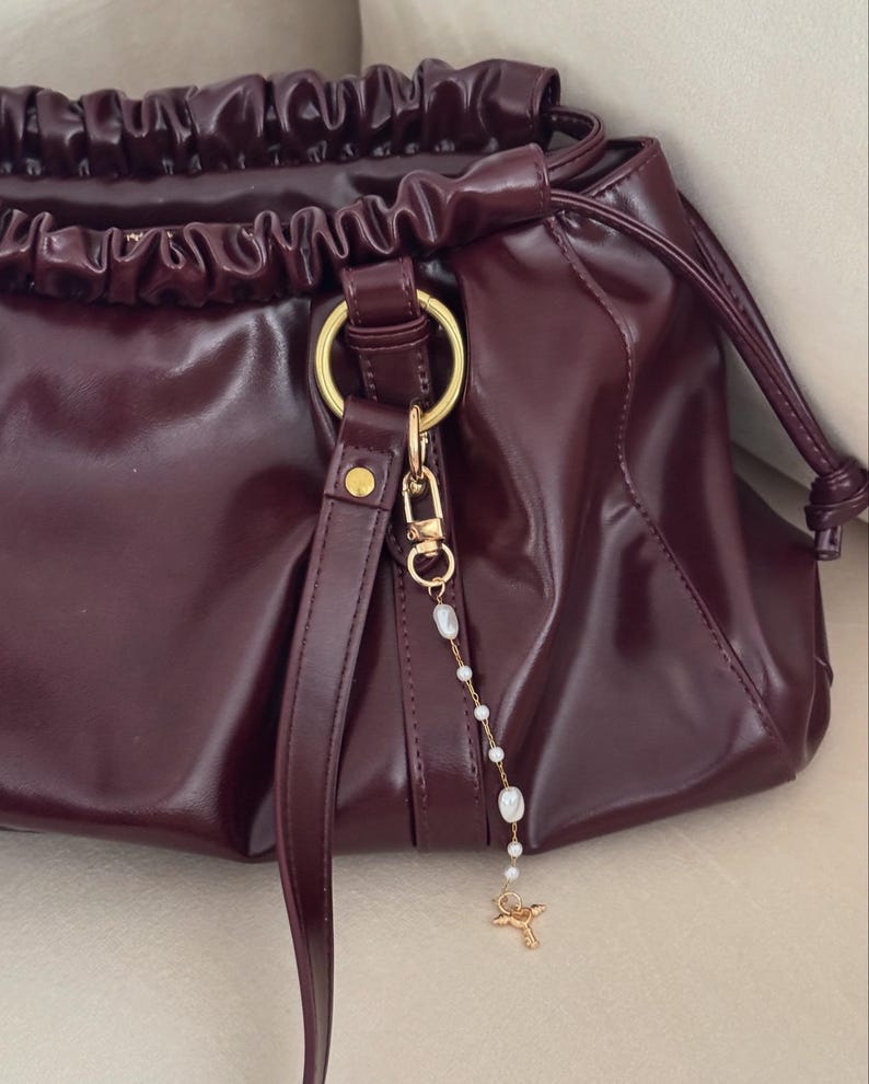 May include: A burgundy leather handbag with a drawstring closure and gold-tone hardware. The bag features a detachable charm with pearls and a small gold-colored figure. The bag has a shiny, smooth finish.