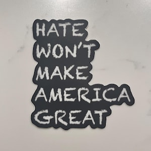 May include: A black sticker with white text that reads "HATE WON'T MAKE AMERICA GREAT". The text is in a handwritten style. The sticker is on a white surface.