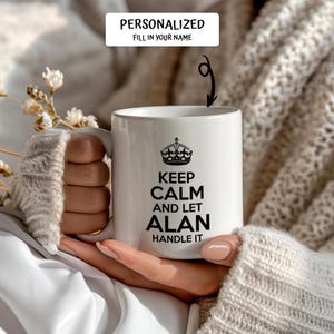 May include: White ceramic mug with the text "KEEP CALM AND LET ALAN HANDLE IT" in black, topped with a crown graphic. The mug is held in a person's hands, with the text "PERSONALIZED FILL IN YOUR NAME" at the top.