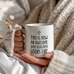 Peut inclure: Mug en céramique blanche avec l'inscription en noir : "THIS IS HOW AN AWESOME GAME DEVELOPER LOOKS LIKE". La tasse a une anse et un symbole de flèche. La tasse est tenue par une personne portant un pull blanc.