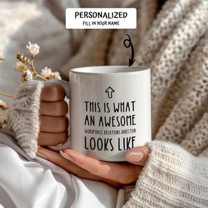 May include: White ceramic mug with the text "THIS IS WHAT AN AWESOME WORKFORCE RELATIONS DIRECTOR LOOKS LIKE" in black. The mug has a personalized section at the top. The mug is held by a person.