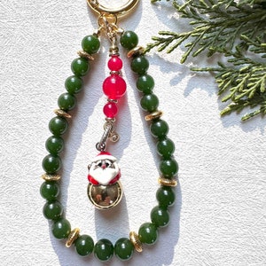 May include: A green jade bead keychain with a gold clasp and a Santa Claus charm. The charm is a bell with a white face and red hat.
