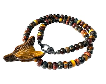 May include: A beaded necklace featuring a carved fox head pendant. The necklace is made of multi-colored tiger eye beads, with a black clasp. The fox head is a light brown color.