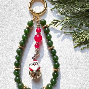 May include: A green jade bead keychain with a gold charm featuring a Santa Claus bell. The keychain also has a red bead and a gold ring.