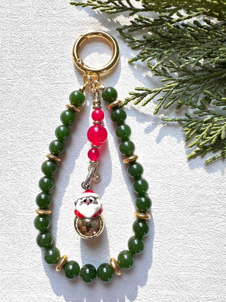 May include: A green jade bead keychain with a gold charm featuring a Santa Claus bell. The keychain also has a red bead and a gold ring.