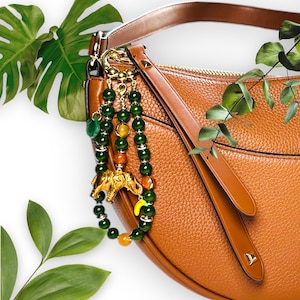 Puede incluir: Un bolso de mano de cuero marrón con un charm de cuentas verdes, naranjas y doradas con una pequeña figura de elefante. El charm está unido a la correa del bolso.