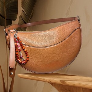 May include: A brown leather crescent-shaped handbag with a beaded strap detail. The bag has a zip closure and a small pocket on the front.