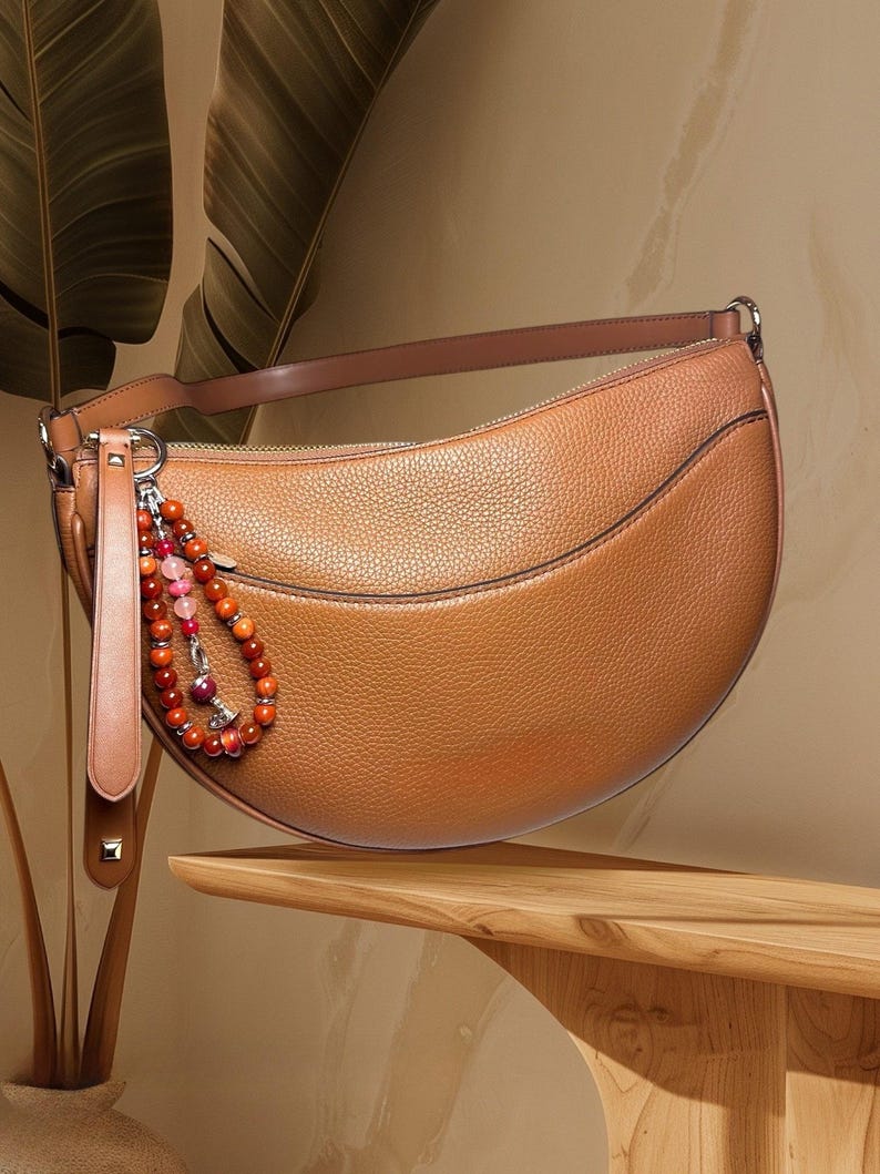 May include: A brown leather crescent-shaped handbag with a beaded strap detail. The bag has a zip closure and a small pocket on the front.