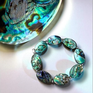 以下が含まれることがあります： アワビの貝殻で作られた、ターコイズ、グリーン、パープルの楕円形のビーズのブレスレット。 イリデッセントビーズは、シルバーカラーのコネクターで繋がれています。 背景には、お揃いのアワビの貝殻があります。
