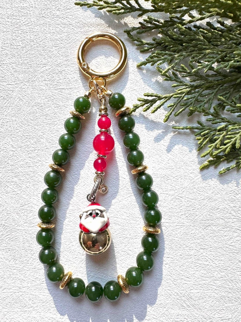 May include: A green jade bead keychain with a gold clasp and a Santa Claus charm. The charm is a bell with a white face and red hat.