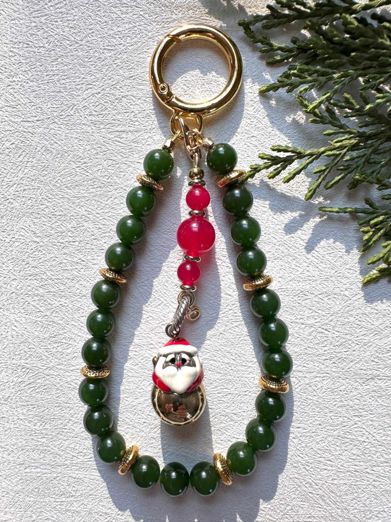 May include: A green jade beaded keychain with a gold clasp and a Santa Claus charm. The charm is a small ceramic figure of Santa with a red hat, white beard, and a gold bell.