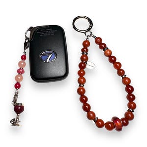 May include: A red and pink beaded keychain with a wine glass charm attached to a black car key fob. The keychain also has a brown beaded loop with a silver charm that says "Lemari".