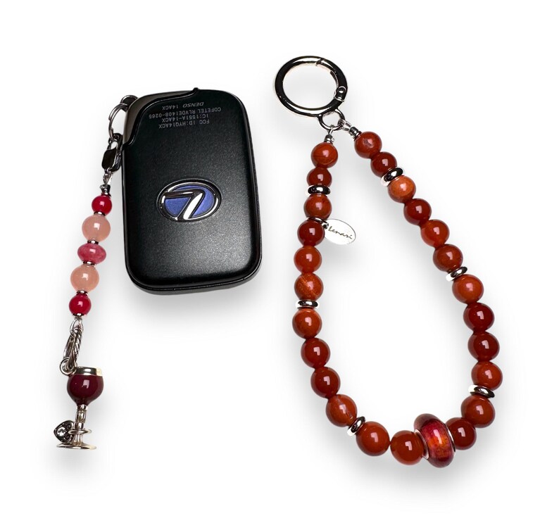 May include: A red and pink beaded keychain with a wine glass charm attached to a black car key fob. The keychain also has a brown beaded loop with a silver charm that says "Lemari".