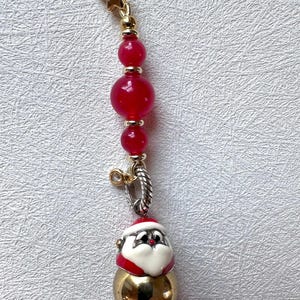 May include: A gold charm with a red and white Santa Claus face on a gold ball. The charm is attached to a chain of red beads with gold spacers.