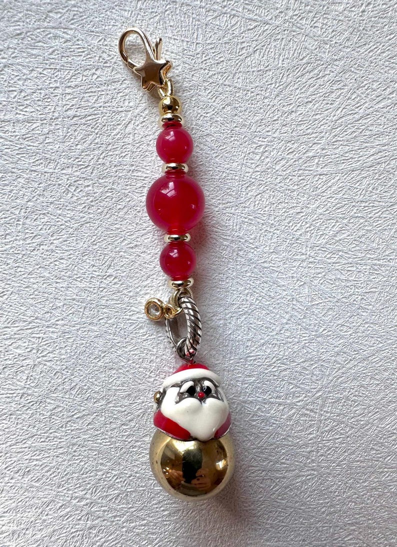 May include: A gold charm with a red and white Santa Claus face on a gold ball. The charm is attached to a chain of red beads with gold spacers.