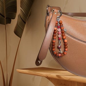 May include: A brown leather handbag with a long strap and a beaded charm. The charm is made of red, pink and orange beads.