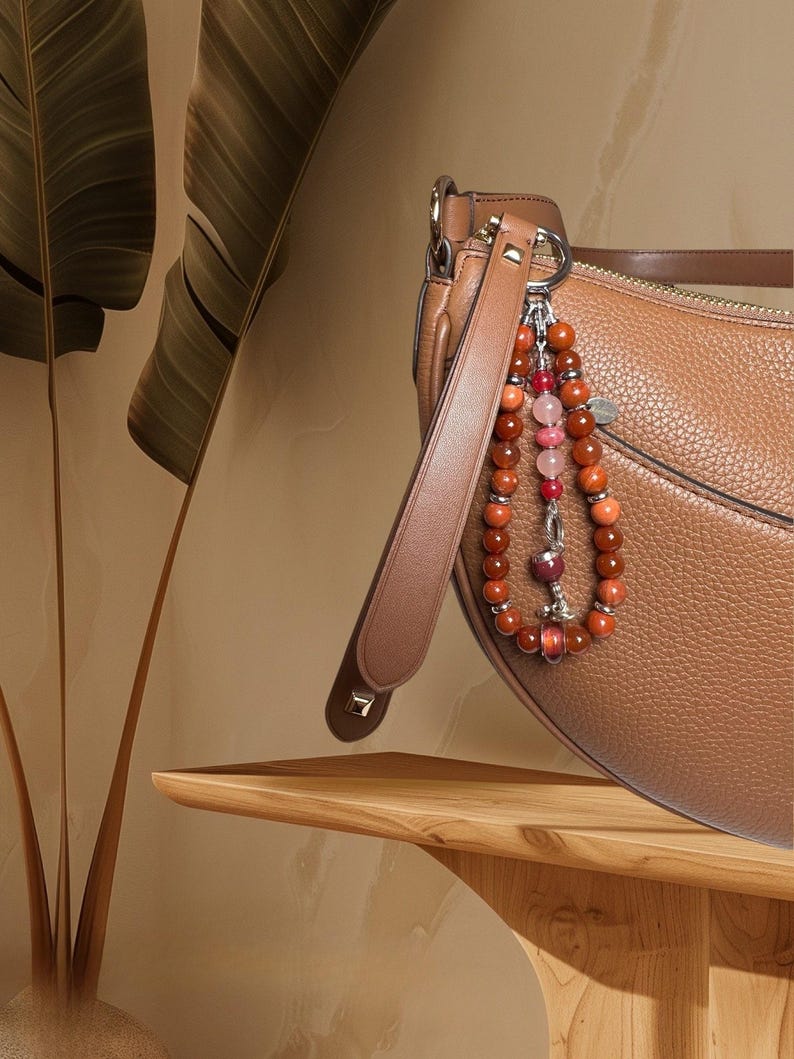 May include: A brown leather handbag with a long strap and a beaded charm. The charm is made of red, pink and orange beads.