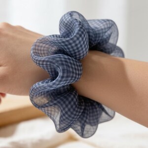 May include: A blue and white gingham scrunchie is worn on a wrist. The fabric is sheer and gathered, creating a voluminous effect. The scrunchie is a hair accessory, adding a touch of style to any hairstyle.