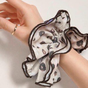 May include: A scrunchie with a beige background and a leopard print pattern. The scrunchie has a brown trim and is worn on a wrist. A gold ring is on a finger.