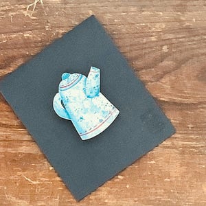 May include: A blue and white ceramic teapot with a red stripe around the top. The teapot is painted with a white and blue speckled pattern.