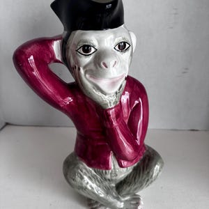 May include: A ceramic monkey figurine, wearing a black hat and a maroon jacket. The monkey has a white face, gray fur, and is posed with its hand to its head. The figurine is a decorative item.