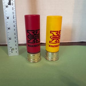 May include: Two shotgun shells, one red and one yellow, with gold-colored bases. Each shell has the text "SUPER" and "WINCHESTER" printed in black. A ruler is placed next to the shells.
