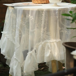 May include: A white lace tablecloth with a ruffled edge. The tablecloth is draped over a round table, with the lace featuring floral patterns. The table is set outdoors, with a basket and a small white dish visible on top.