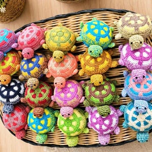 May include: A collection of small, multi-colored crocheted turtle toys arranged in a woven basket. Each turtle features a distinct color combination on its shell and body, including shades of blue, pink, yellow, green, purple, and brown.