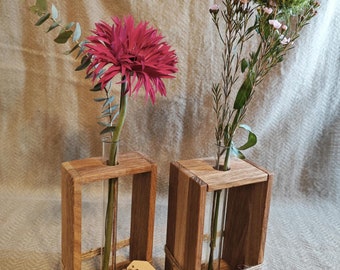 May include: Two wooden flower vases with glass test tubes for holding flowers. The vases are made of natural wood and have a rustic look. One vase holds a pink gerbera daisy and the other holds a bouquet of small white and green flowers. The vases are tied with twine and have a tag that says "Mit Liebe Handgemacht".