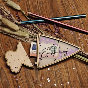 May include: A wooden gift box with the text "zur Einschulung" and a butterfly-shaped wooden cutout. The box contains money and is decorated with purple confetti and pencils.