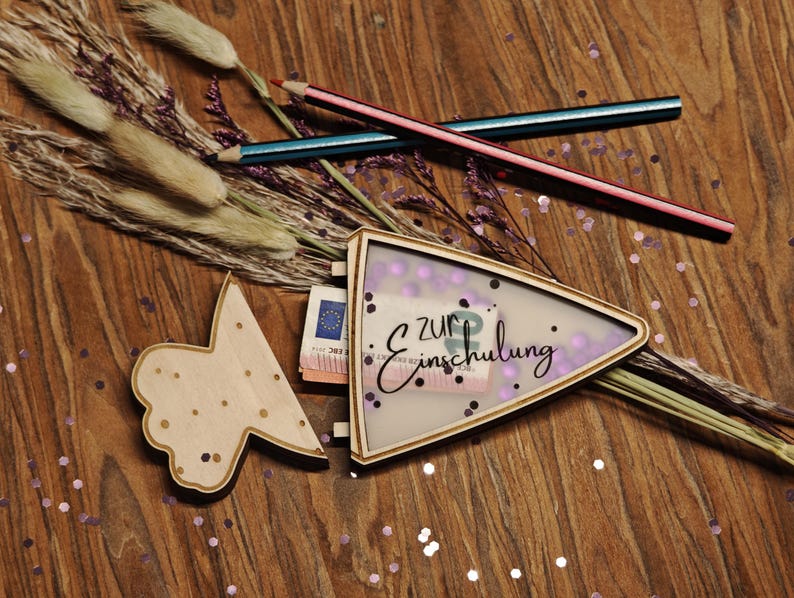 May include: A wooden gift box with the text "zur Einschulung" and a butterfly-shaped wooden cutout. The box contains money and is decorated with purple confetti and pencils.