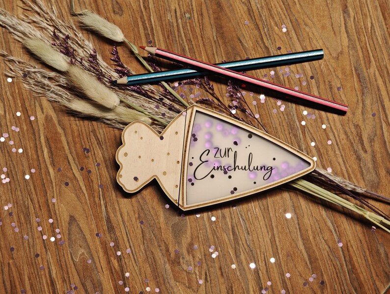 May include: A wooden school cone-shaped decoration with the text "zur Einschulung" in black script, filled with purple confetti. Two pencils and dried plants are arranged on a wooden surface scattered with glitter.