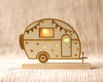 May include: A wooden camper figurine with a light inside. The camper is white with brown trim and has a heart-shaped door. The camper is decorated with small hearts and bunting.