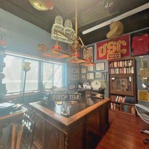 May include: An office space with a large wooden desk, a vintage-style chandelier, and a sign that reads "BIRCH TEKR". The walls are decorated with framed items, a USC banner, and a bookshelf. The room has a vintage aesthetic.