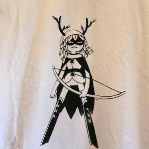 May include: White t-shirt featuring a black and white illustration of a character with antlers, a mask, and a bow and arrow. The character is wearing a cape and leggings with star accents. The design is a graphic print.