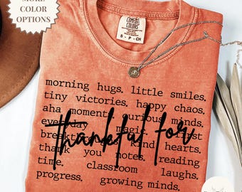 May include: A burnt orange crew neck t-shirt with a distressed texture. The shirt features black text listing phrases like morning hugs, little smiles, tiny victories, happy chaos, and the script word thankful for in the center.