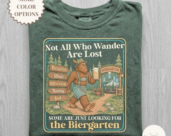 May include: A moss green t-shirt featuring a graphic of a brown Bigfoot wearing lederhosen and a hat, holding a beer mug. The text reads: Not All Who Wander Are Lost, Some Are Just Looking for the Biergarten, with signs for Biergarten, Music, Pretzels, Dancing, and Food.