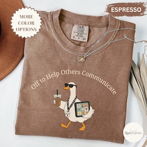 May include: An espresso-colored t-shirt featuring a graphic of a white duck wearing sunglasses, holding a coffee cup, and carrying a tablet with communication icons. The text reads Off to Help Others Communicate. A silver necklace rests on the collar.